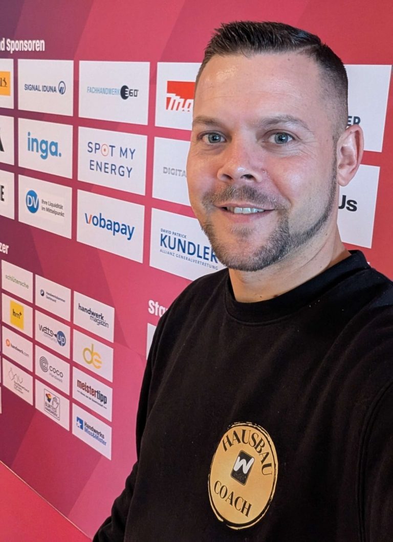 David John Walters Ein Mann mit kurzem Haar lächelt vor einer Wand mit Sponsorenlogos.